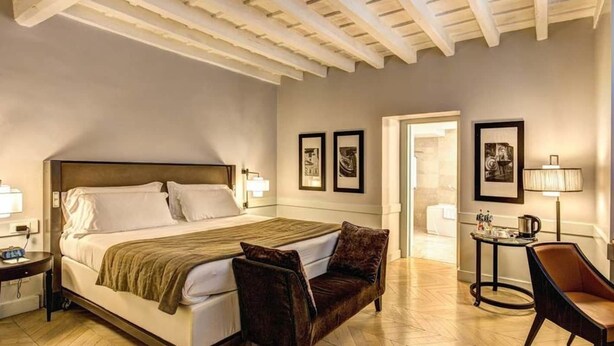 A hotel room with a large bed, a small sofa, a desk with a lamp, and framed pictures on the wall. - Suite Giulia 1 (Rome)