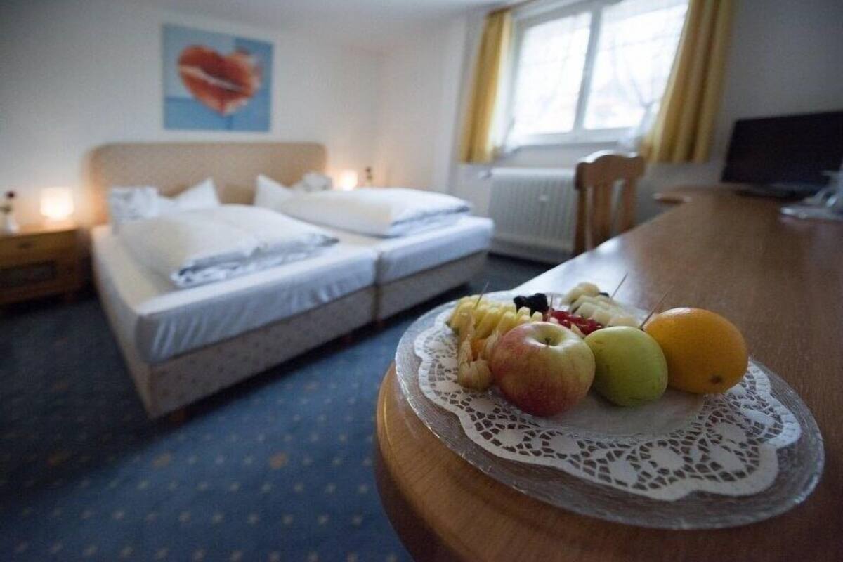 Habitación de hotel con cama doble, mesita de noche con una bandeja de fruta, lámpara de noche y una ventana con cortinas.