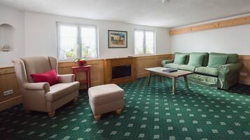 Un salon avec un tapis à motifs verts, un fauteuil beige agrémenté d’un coussin rouge, un canapé vert, une table basse en bois et une cheminée.