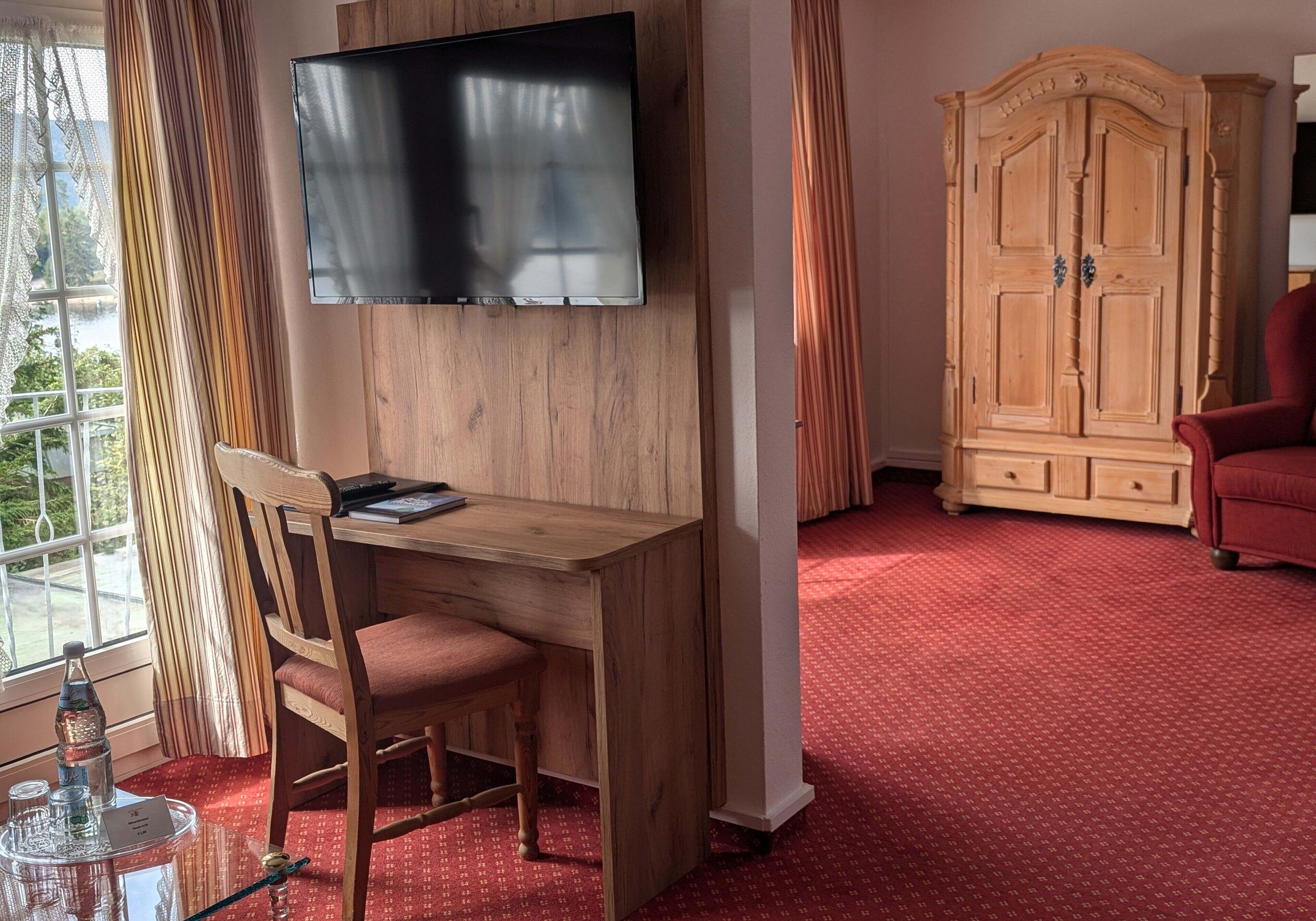 A room with a wooden desk, a television mounted on the wall, a red carpeted floor, a wardrobe, and a red armchair.