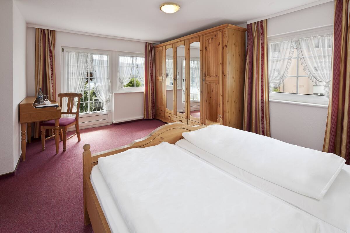 Un dormitorio con una cama de madera, un escritorio y una silla. Hay ventanas con cortinas, y vistas al exterior.