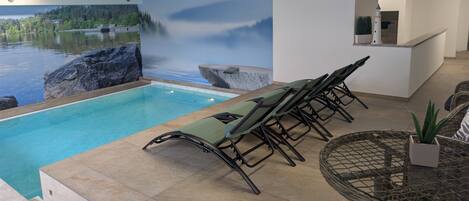 A pool area with a view of a lake and waterfall mural, a lounge with chairs, and a small table with a plant.