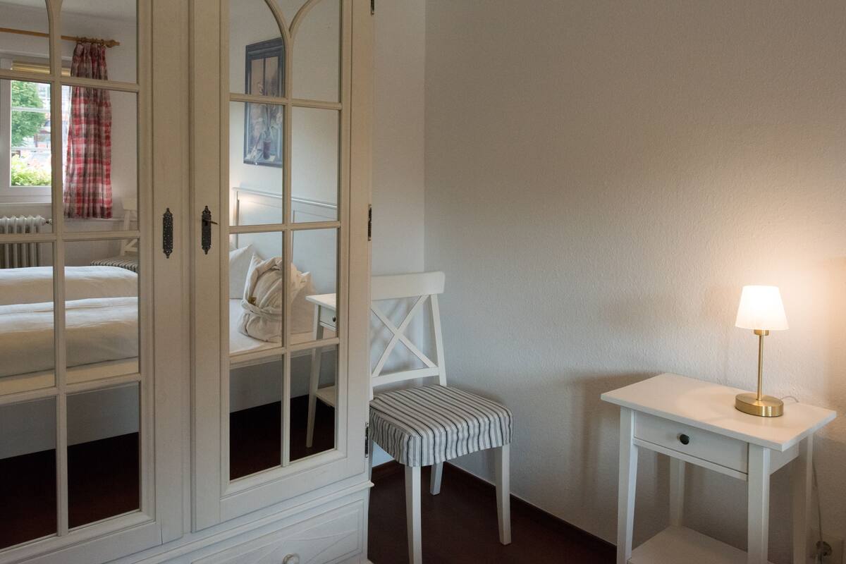 Una habitación con cama, ventana con vistas, una silla, mesita de noche y lámpara.