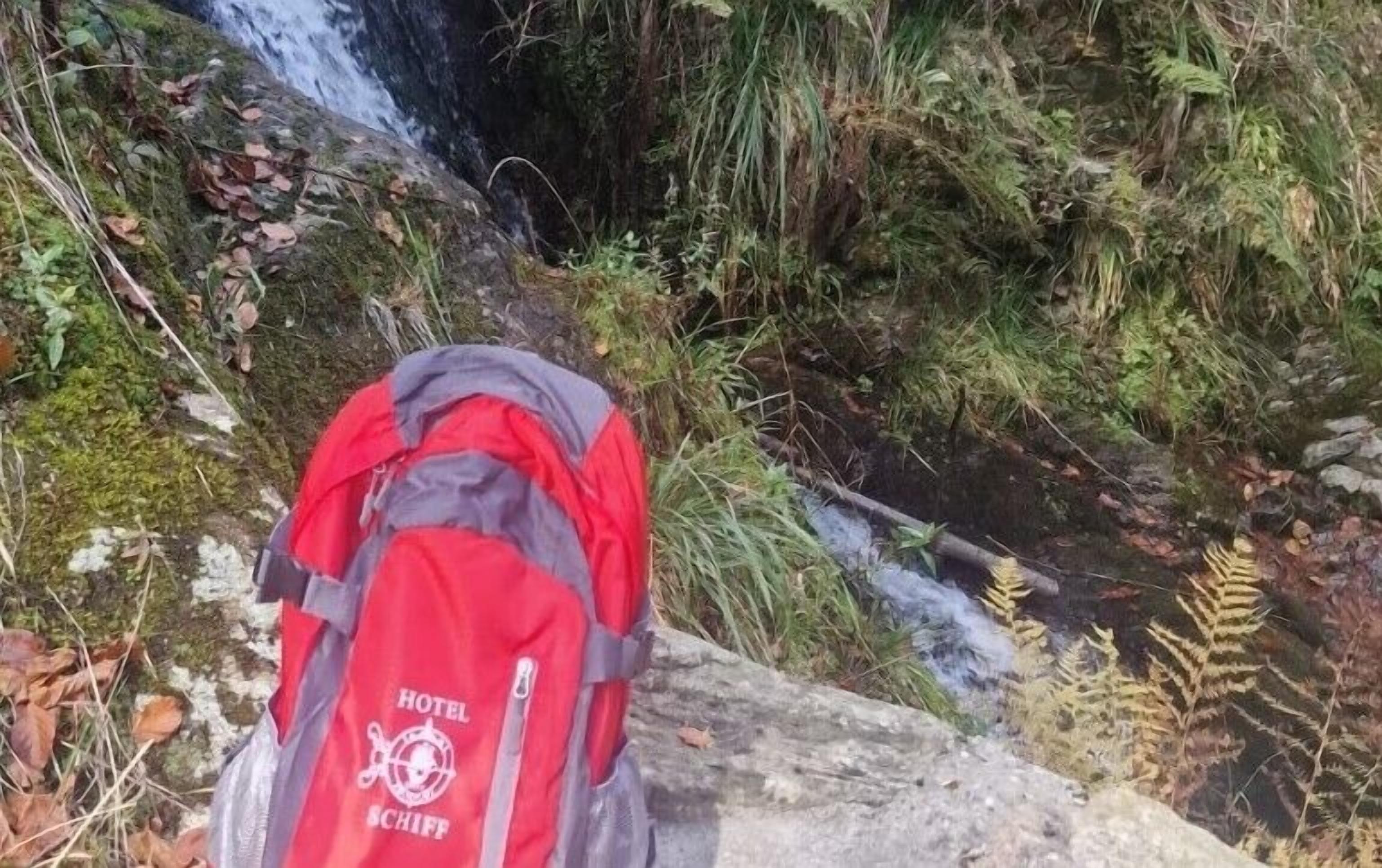 A red backpack with "HOTEL SCHIFF" on it, placed on a rock near a waterfall.