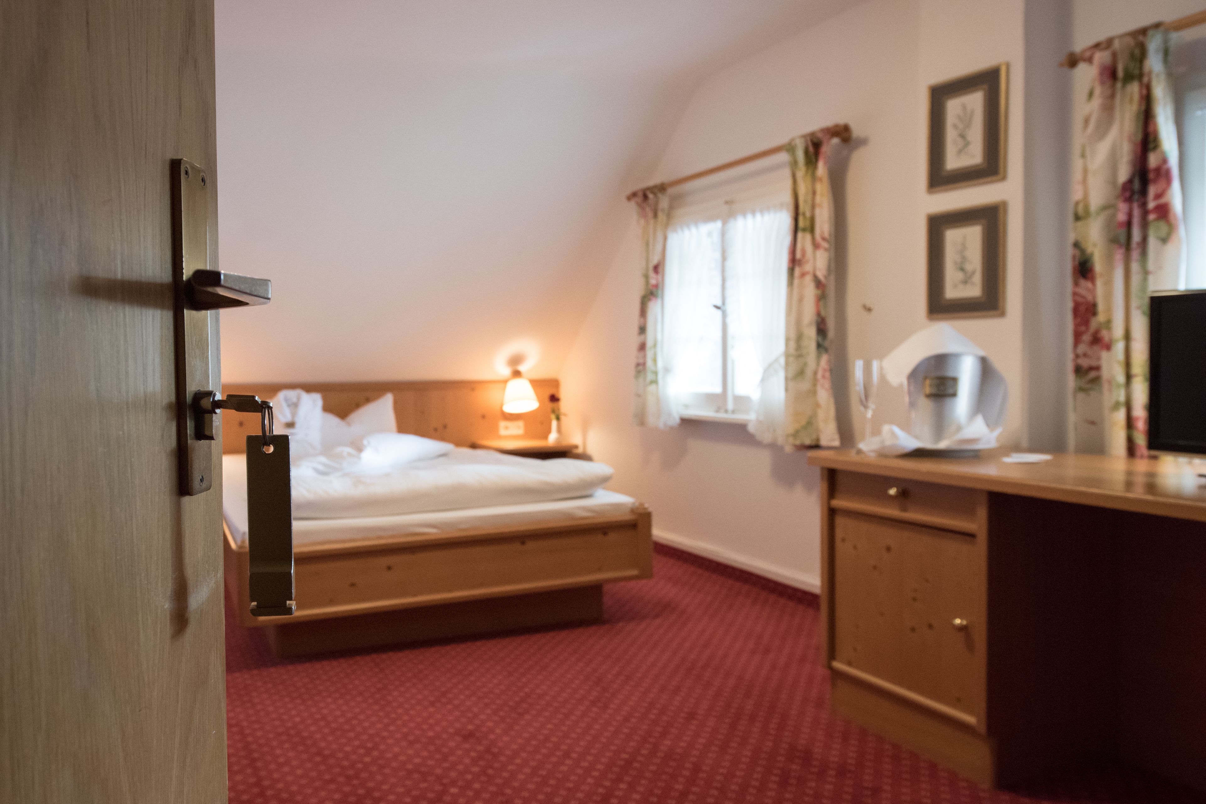 A hotel room with a bed, bedside table, lamp, window with curtains, and a desk with a chair.