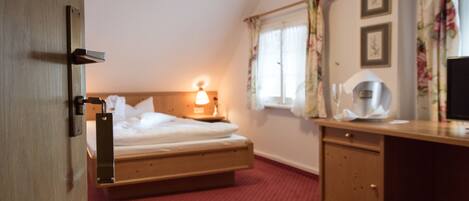 A hotel room with a bed, bedside table, lamp, window with curtains, and a desk with a chair.