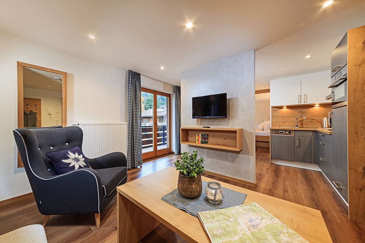 A modern living room with a wooden dining table, a comfortable armchair, a TV mounted on the wall, and a kitchen area with grey cabinets and wooden countertops.