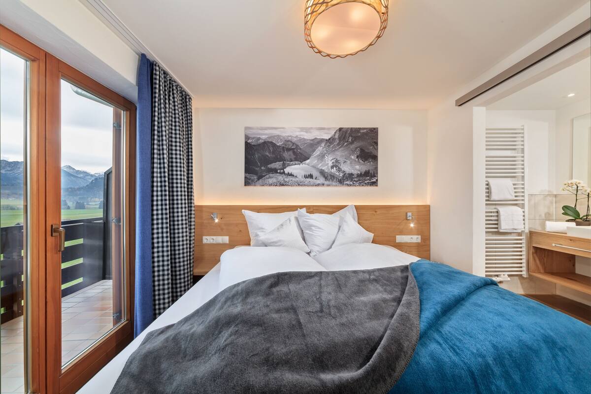 A bedroom with a bed, a view of mountains through a sliding door, a painting on the wall, and a bathroom with a towel rack.