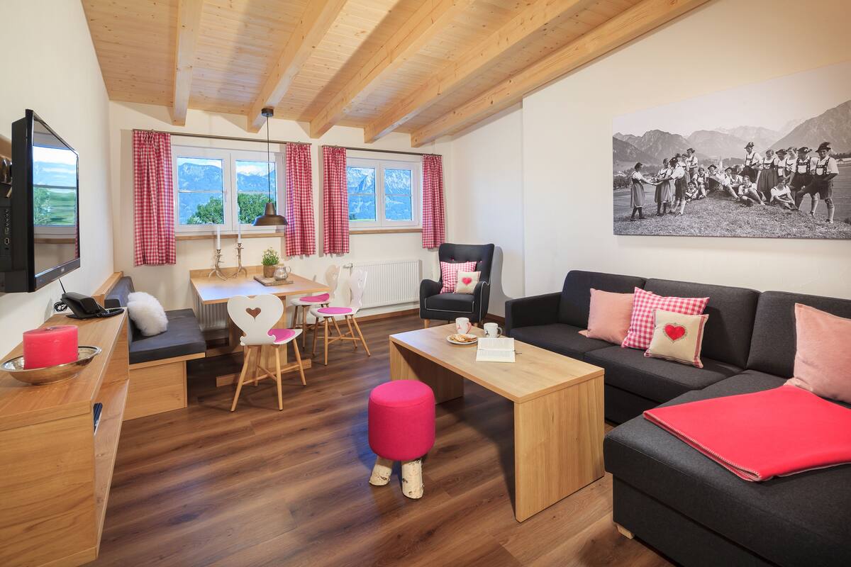A cozy living room with a wooden ceiling, a black sofa with pink cushions, a wooden coffee table, and a red ottoman.