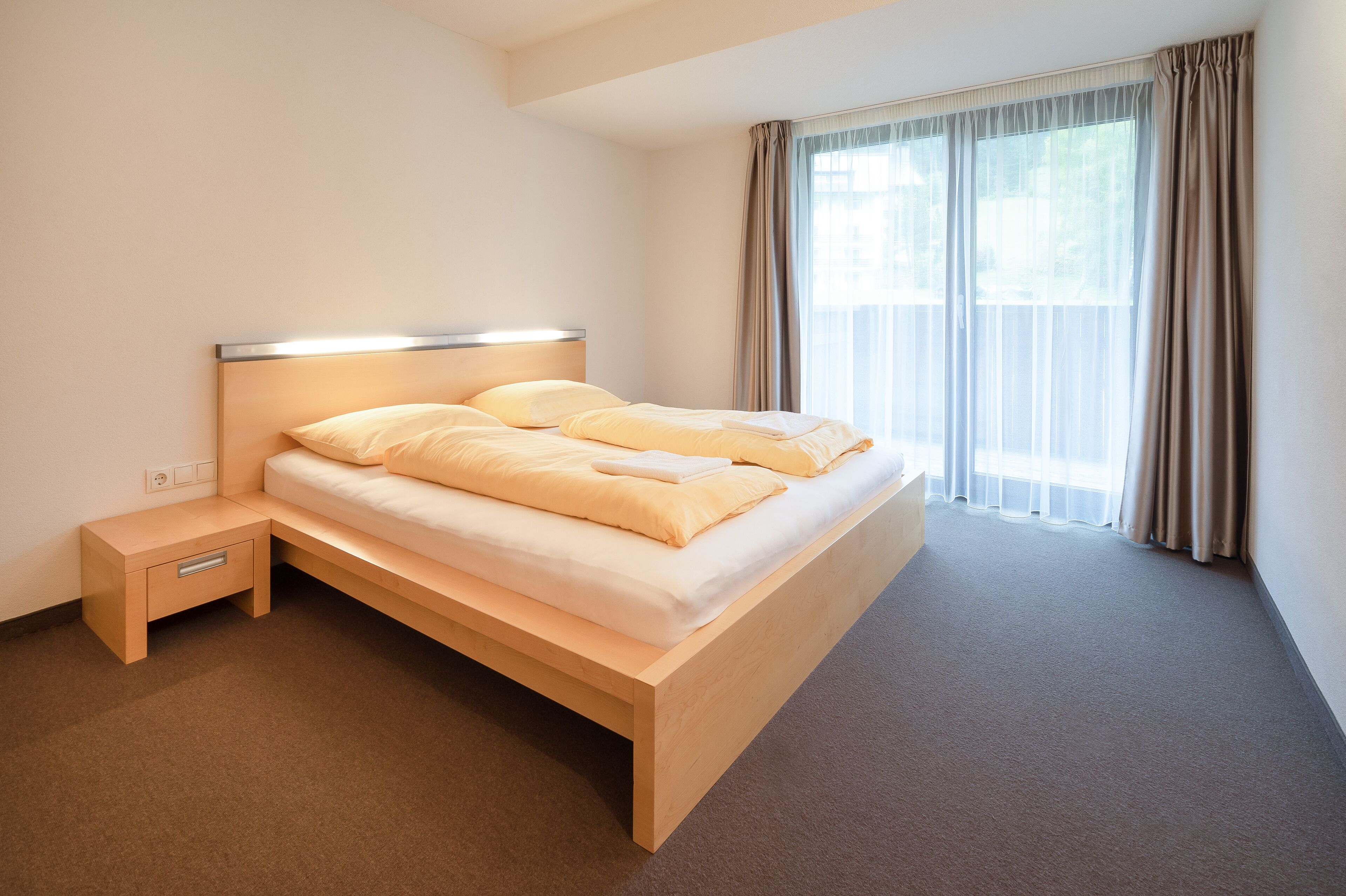 A modern hotel room with a wooden bed, a bedside table, and a large window with curtains.