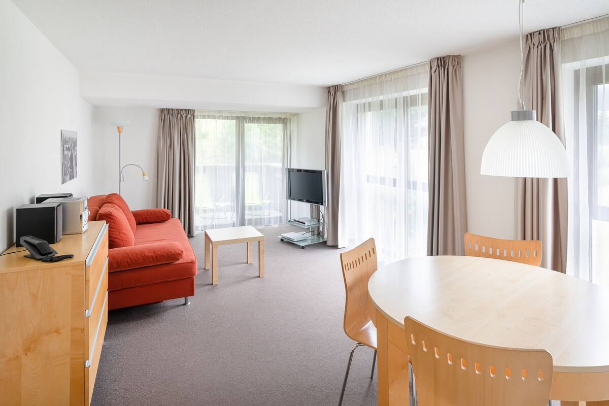 A hotel room with a wooden desk, a red sofa, a dining table, and a TV.