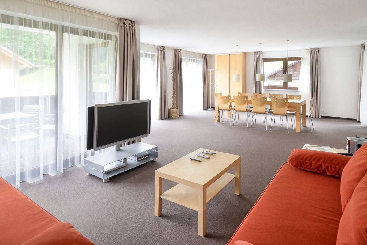 A modern living room with a flat-screen TV, a wooden coffee table, and an orange sofa. There is a dining area with yellow chairs and a table in the background.