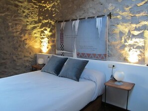 Un dormitorio con pared de piedra, cama con ropa de cama blanca, dos almohadas y mesita de noche con lámpara.