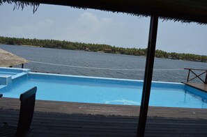 A swimming pool with a wooden deck and a view of a body of water and a distant shoreline with buildings. - Villa Assinie Bord de Lagune (Assinie)