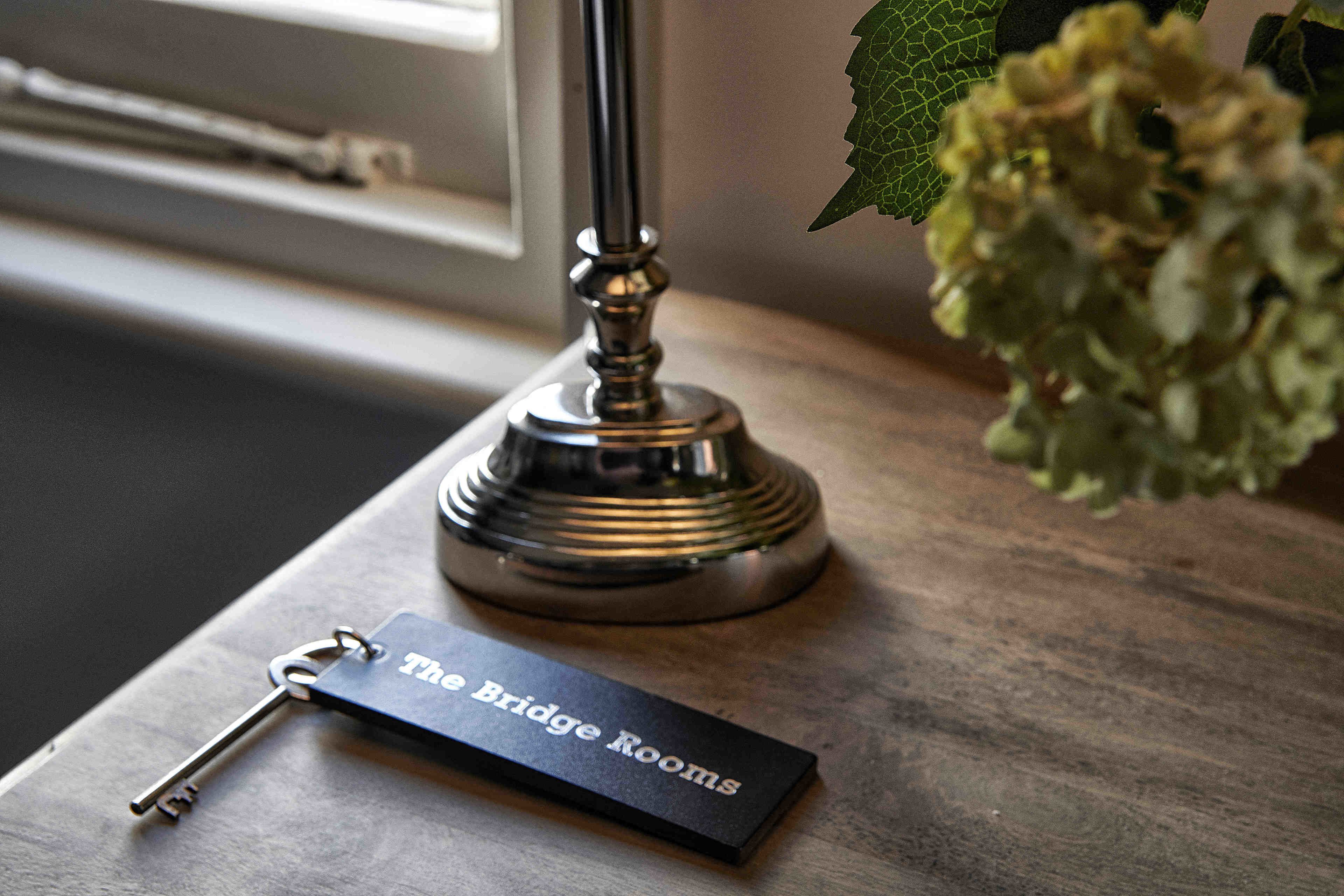 A key and a nameplate on a wooden surface with a lamp and a plant in the background.