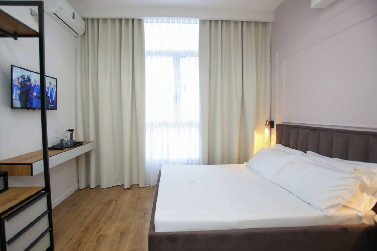 Habitación de hotel con cama, escritorio, televisor y ventana con cortinas.
