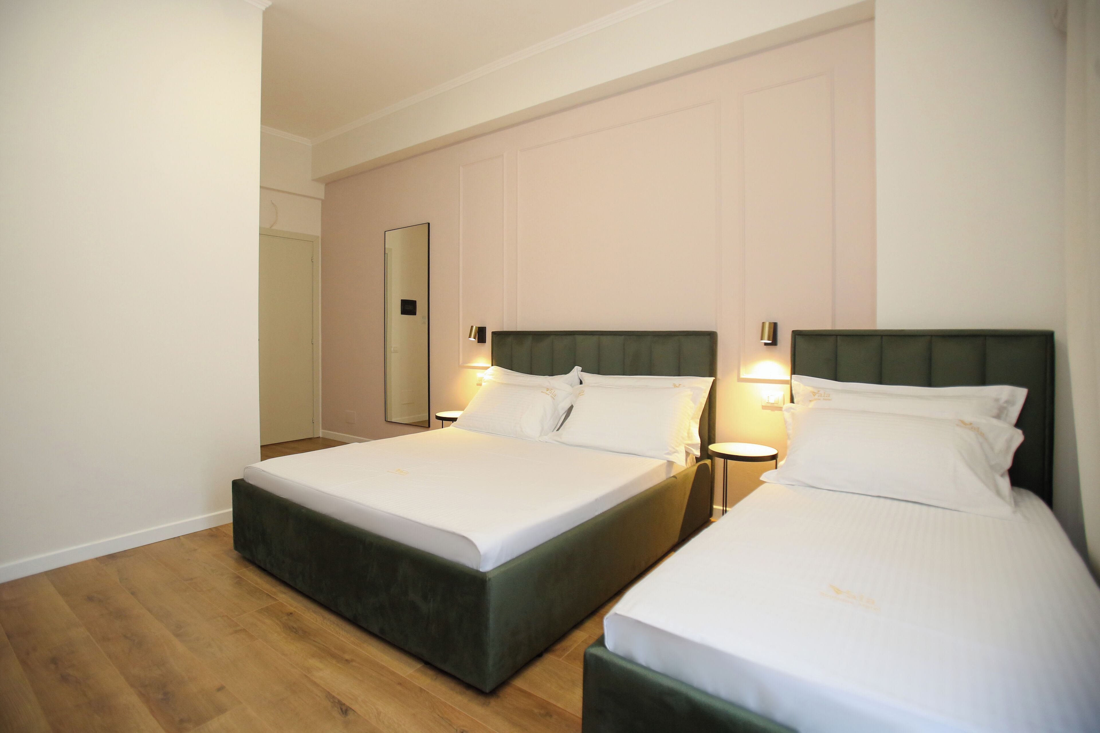 A hotel room with two beds, each with a green headboard and white bedding. There are bedside lamps and a mirror on the wall.