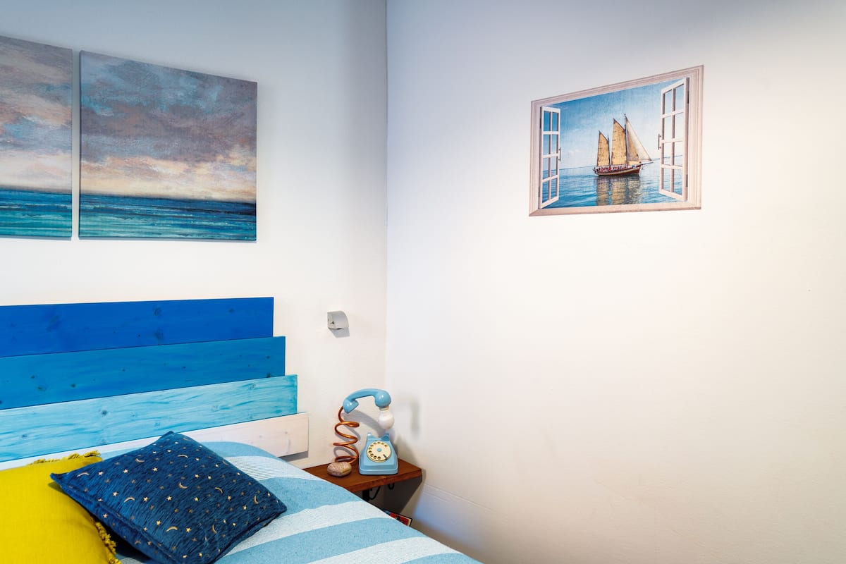 Un dormitorio con cama, mesita de noche, teléfono azul y una obra de arte en la pared que muestra un velero.