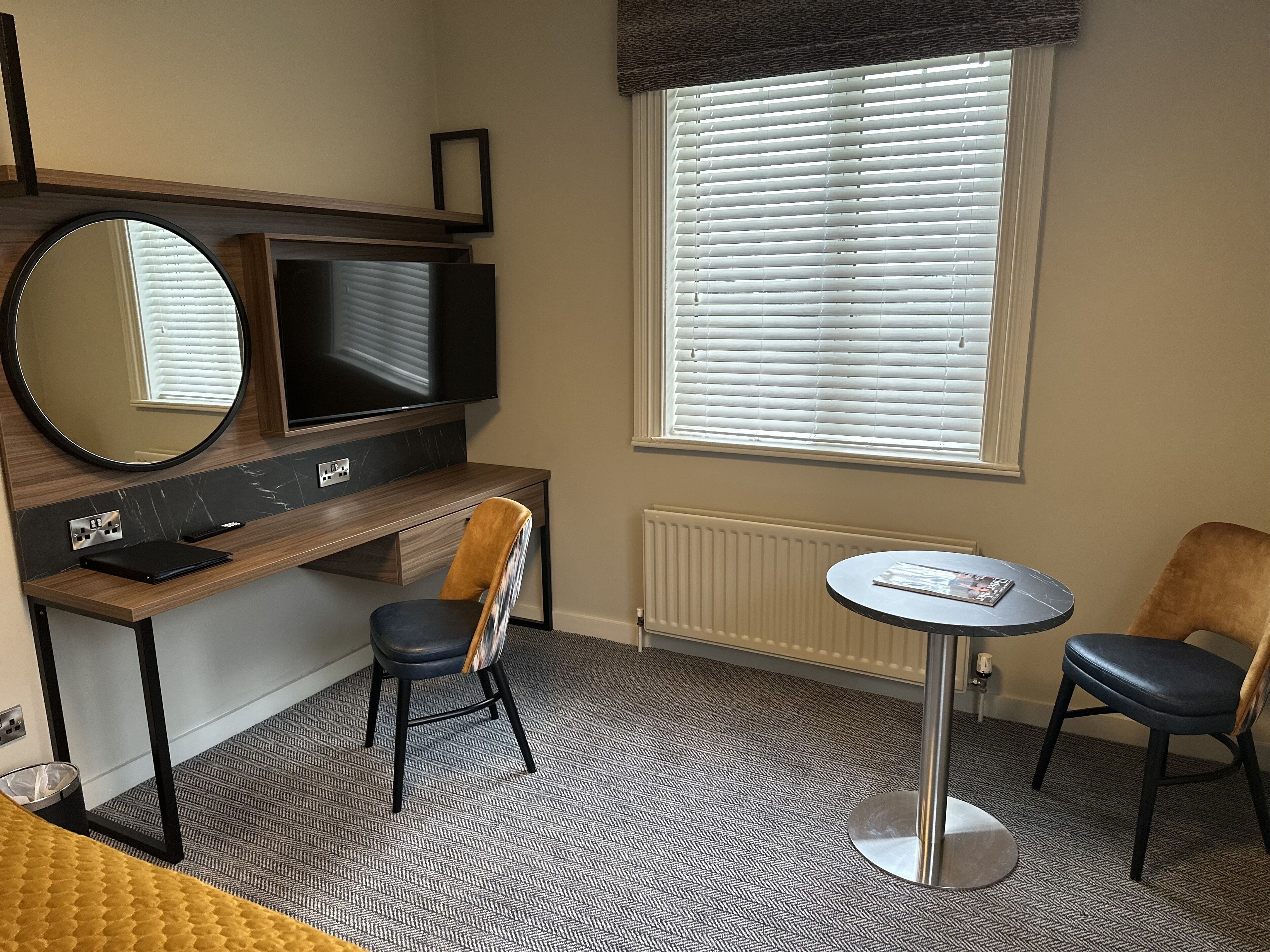 Ett hotellrum med ett skrivbord i trä, en stor spegel, en tv, två stolar, ett litet runt bord och ett fönster med persienner.