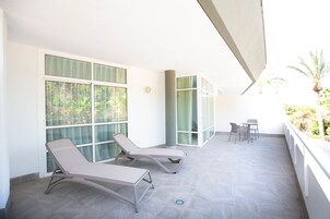 Un espacio al aire libre iluminado por el sol, con dos sillones de descanso y una mesita, ventanas amplias con cortinas, y vistas a zonas verdes.