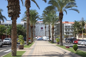 Entrada de hotel con palmeras, cielo despejado y coches aparcados.