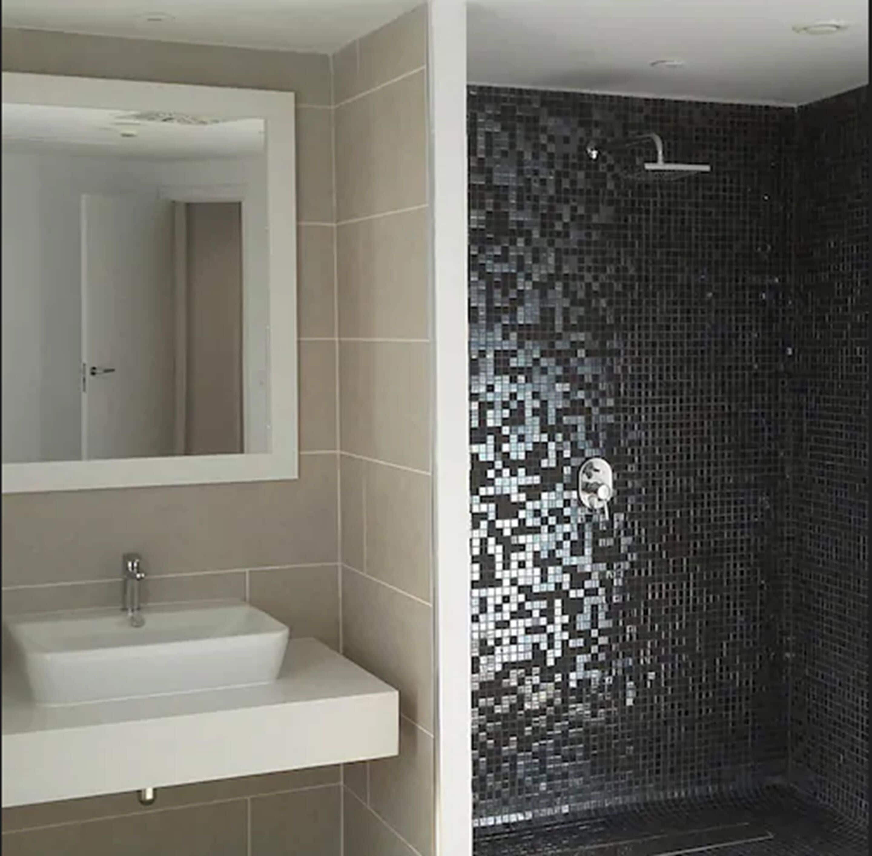 Une salle de bain avec un lavabo blanc, un miroir et une douche dont le mur est recouvert de mosaïques.