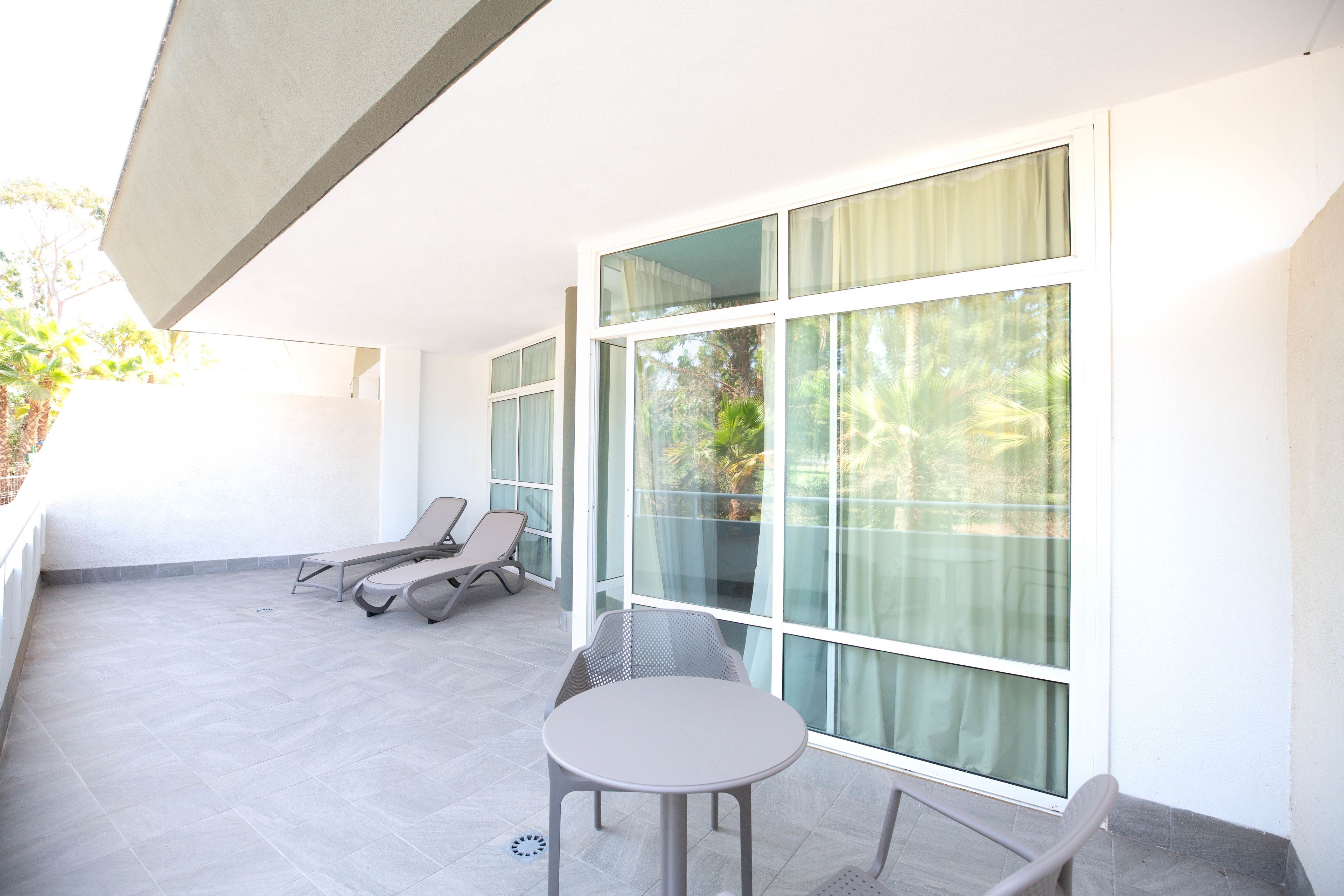 A sunlit balcony with a table and two lounge chairs, large windows, and a view of greenery.