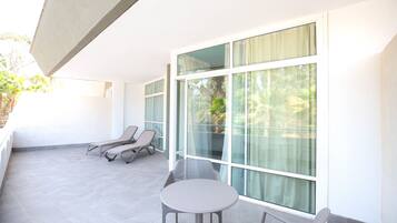 A sunlit balcony with a table and two lounge chairs, large windows, and a view of greenery.