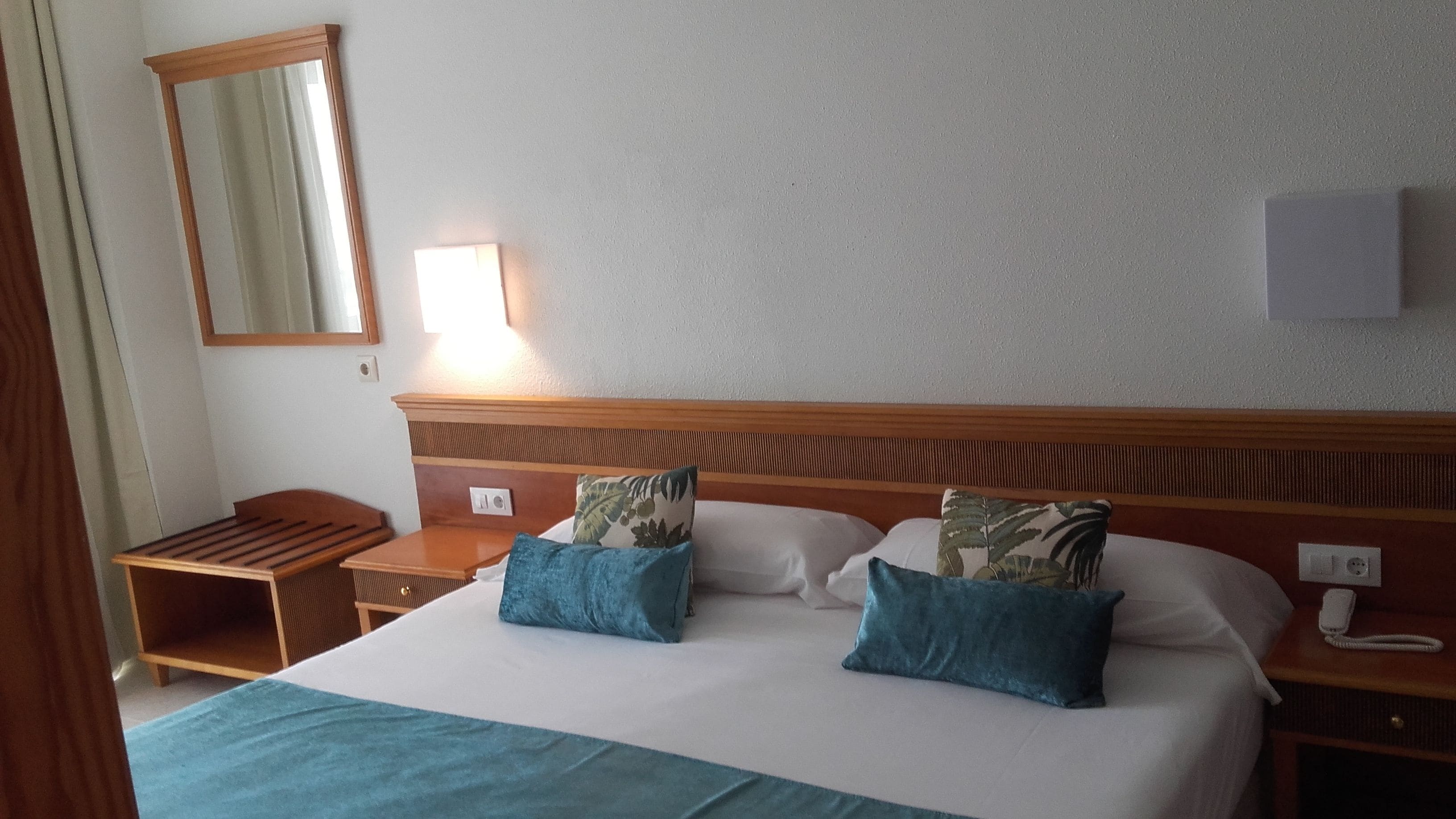 Ein Hotelzimmer mit einem Bett, zwei Kissen, einem Nachttisch, einer Lampe, einem Spiegel und einem Telefon.
