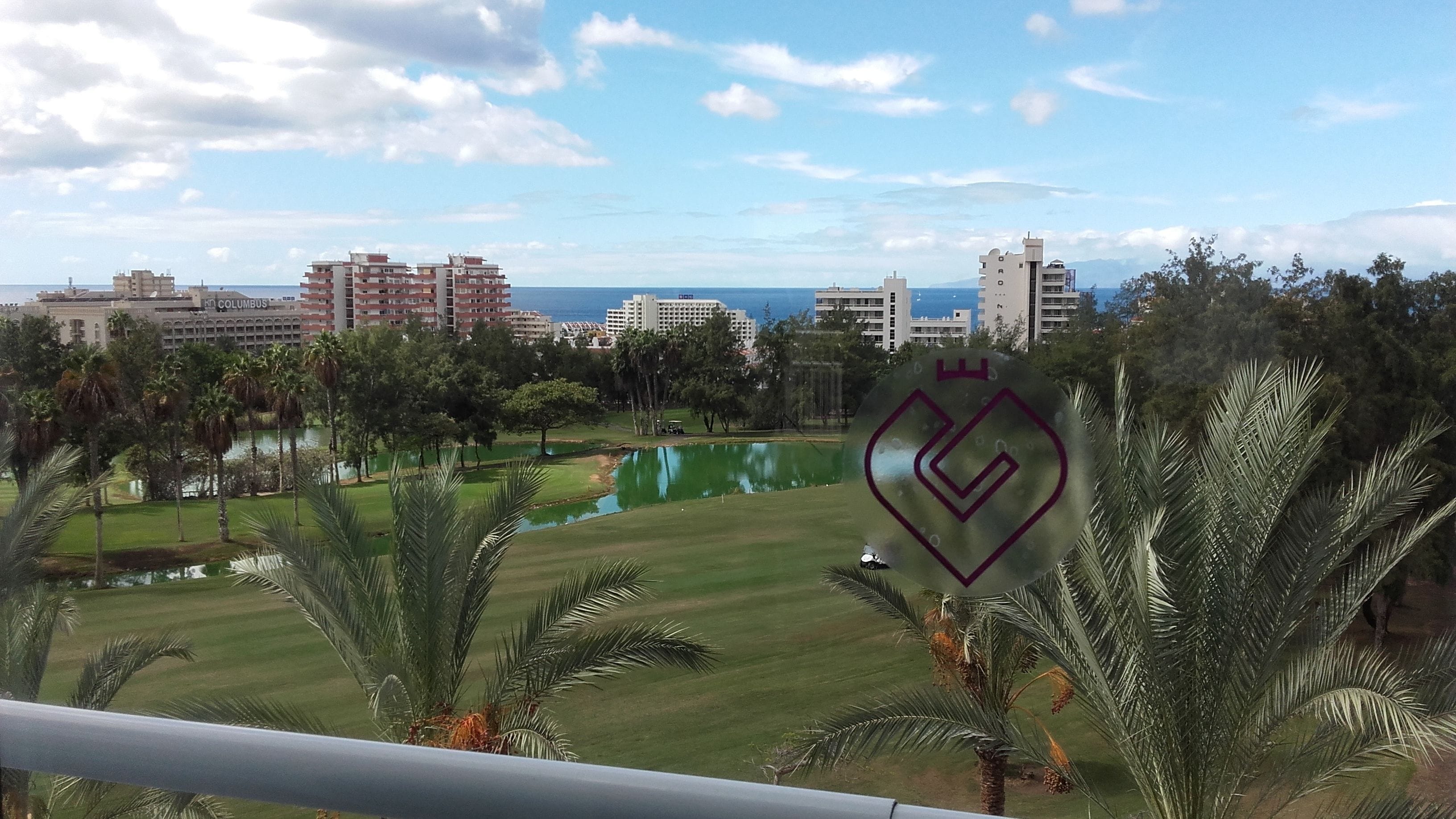 Blick auf einen Golfplatz mit Teich, Palmen und Gebäuden im Hintergrund.