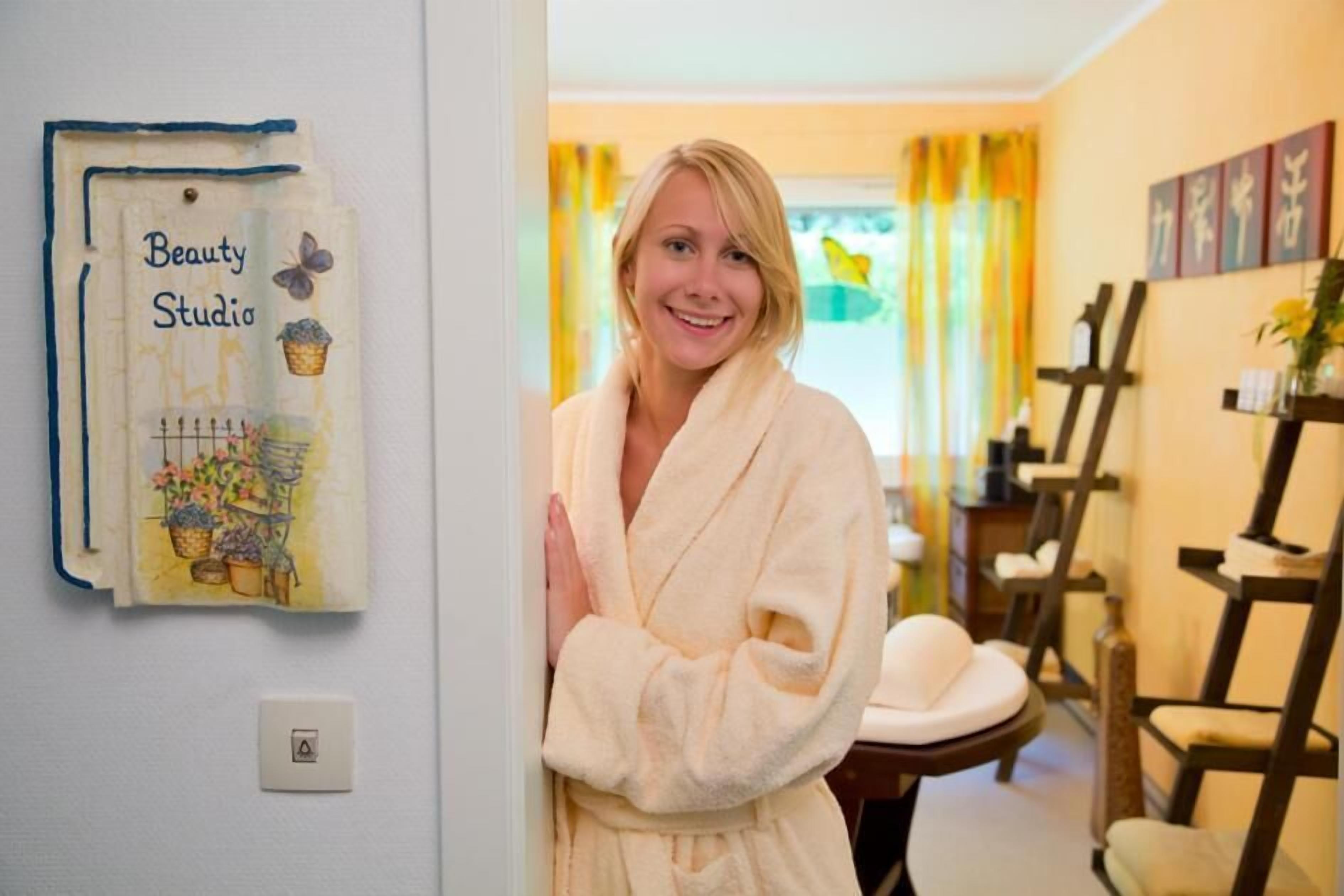 A person in a robe standing in a room with a wall-mounted sign reading "Beauty Studio," a chair, and a table.