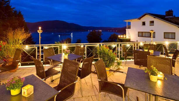 An outdoor terrace with tables and chairs, overlooking a body of water and hills at dusk. - Waldhotel Wiesemann (Waldeck)