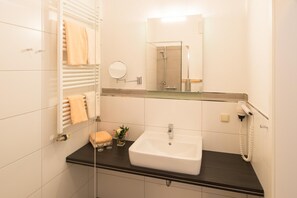 Ein Badezimmer mit einem weißen Waschbecken, einer Duschkabine aus Glas, einem Handtuchhalter mit Handtüchern, einer kleinen Vase mit Blumen und einem an der Wand befestigten Föhn.