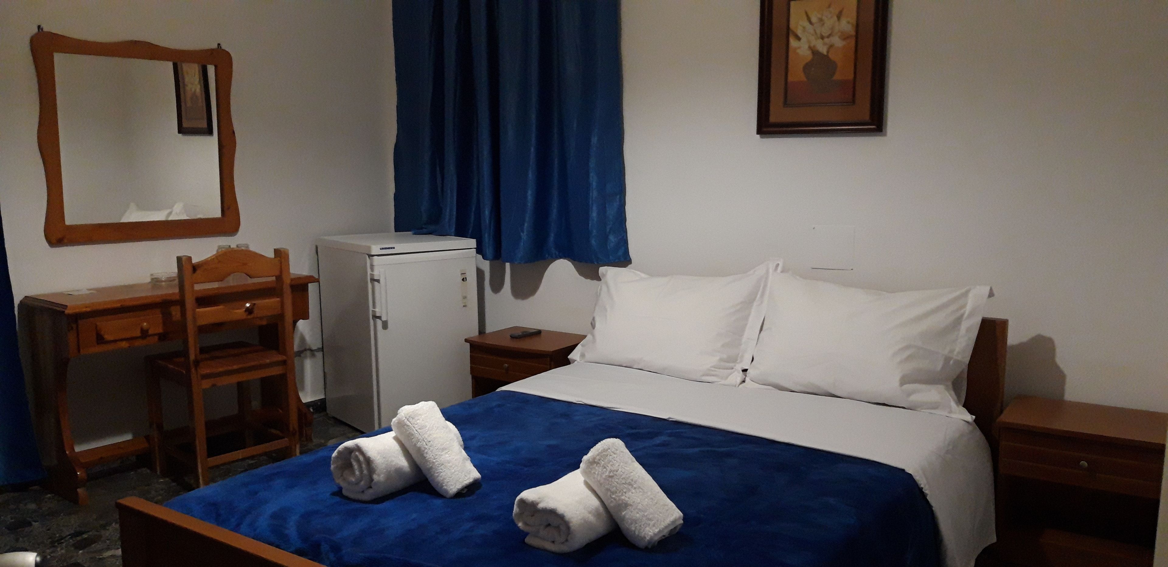 A hotel room with a bed, a desk, a chair, a small refrigerator, and a blue curtain.