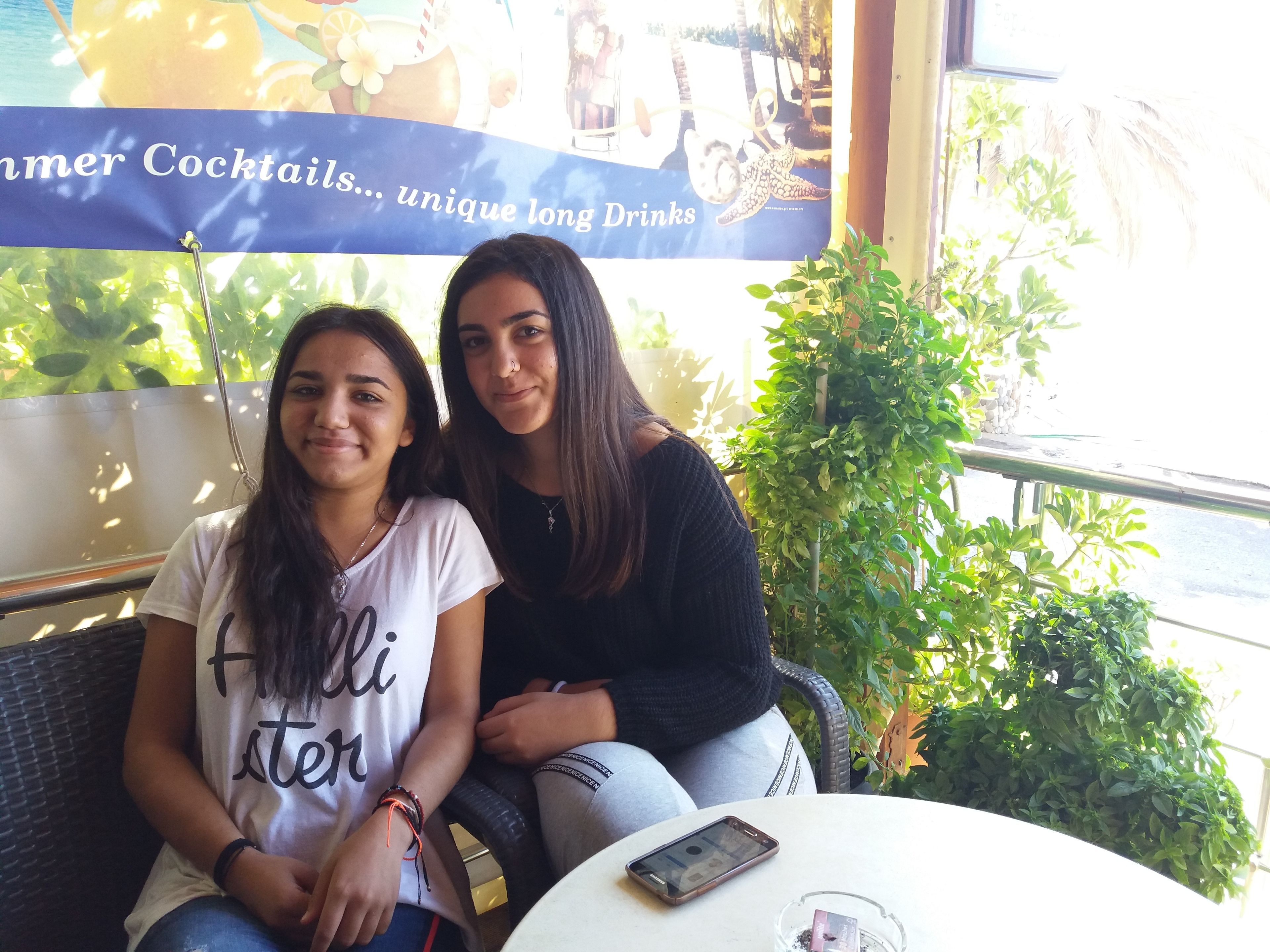 Dos personas sentadas en sillas de mimbre junto a una mesa, con un teléfono inteligente y un vaso de bebida. Se observa una pancarta con texto al fondo.