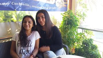 Dos personas sentadas en sillas de mimbre junto a una mesa, con un teléfono inteligente y un vaso de bebida. Se observa una pancarta con texto al fondo.