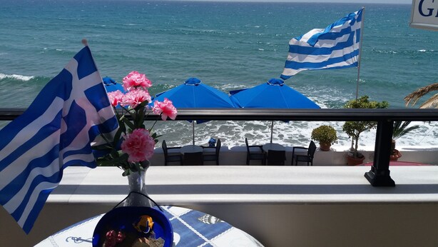 Un balcón con vista al mar, con banderas azul y blanco a rayas, flores rosadas y mobiliario de exterior.
