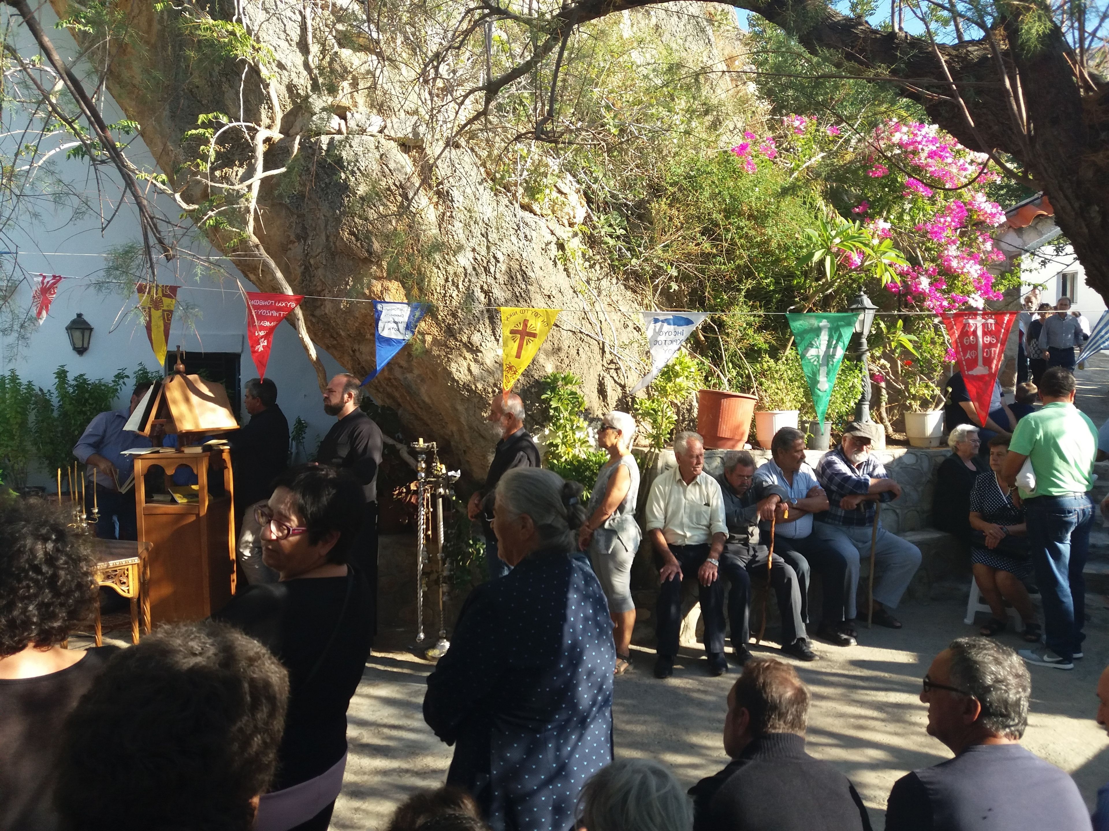 A group of people gathered outdoors, some standing and some seated, with a large tree and potted plants in the background.