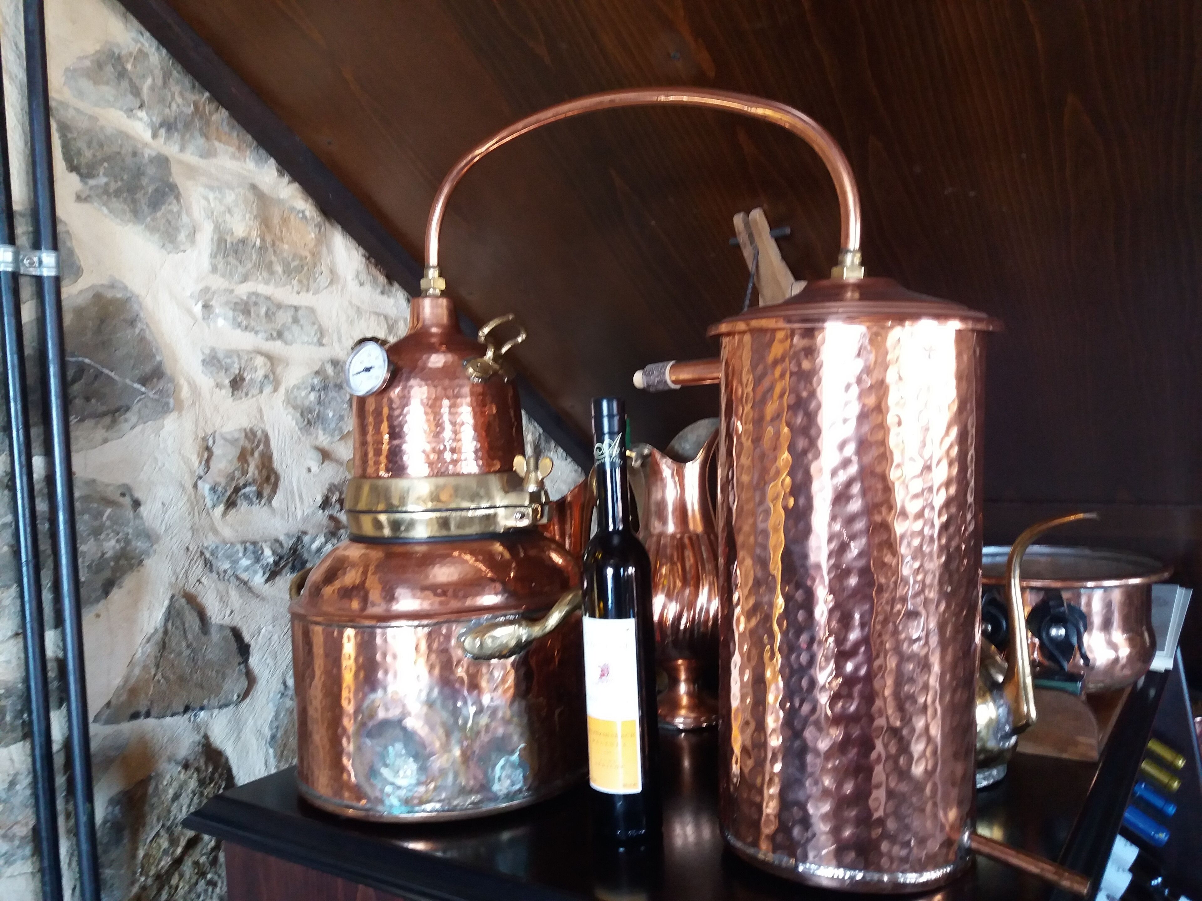 A copper distillation apparatus and a bottle of wine on a wooden table against a stone wall.