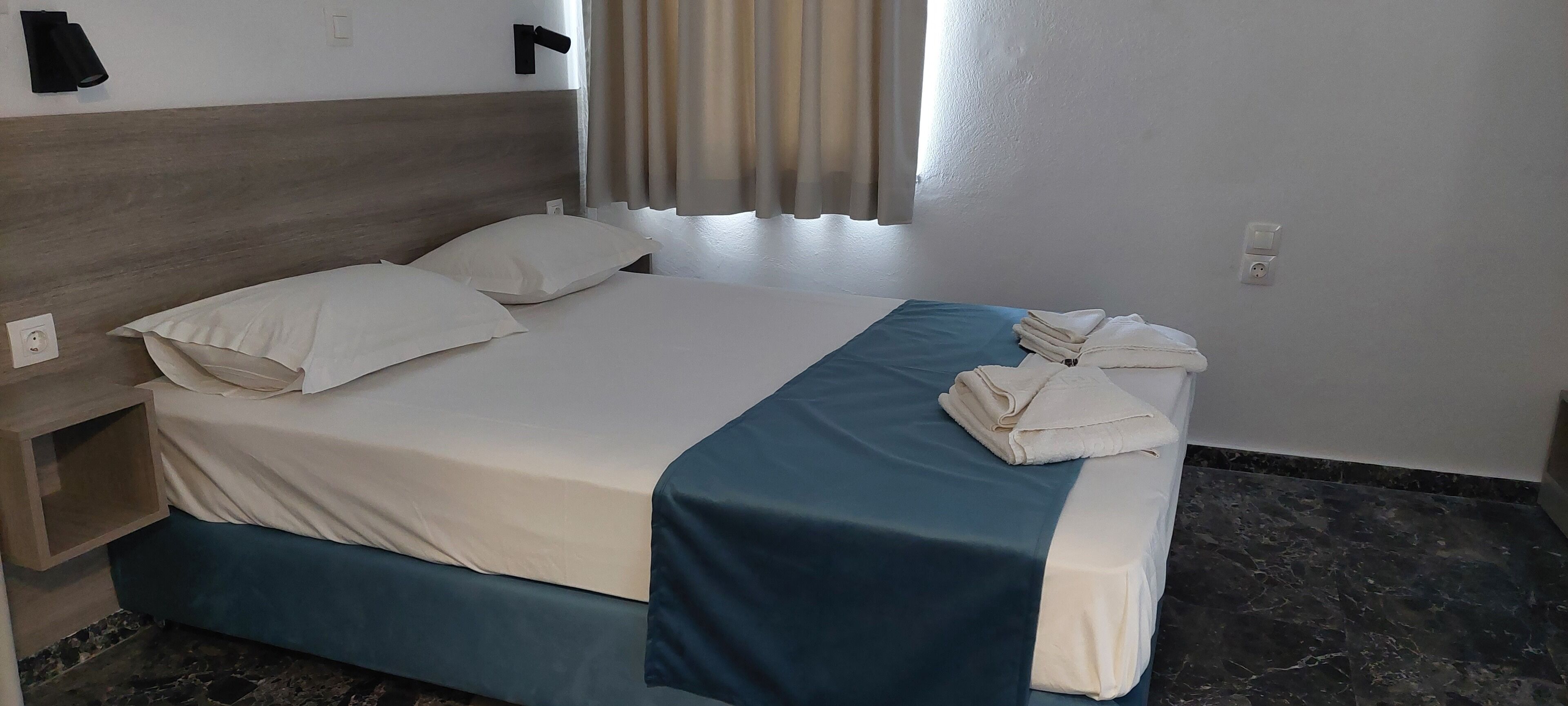 A neatly made bed with white linens and a blue accent, a wooden headboard, and a window with curtains.