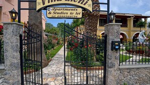 Une entrée fermée par un portail mène à la villa Eleni, et une pancarte indique qu’elle propose des appartements et des studios à louer.