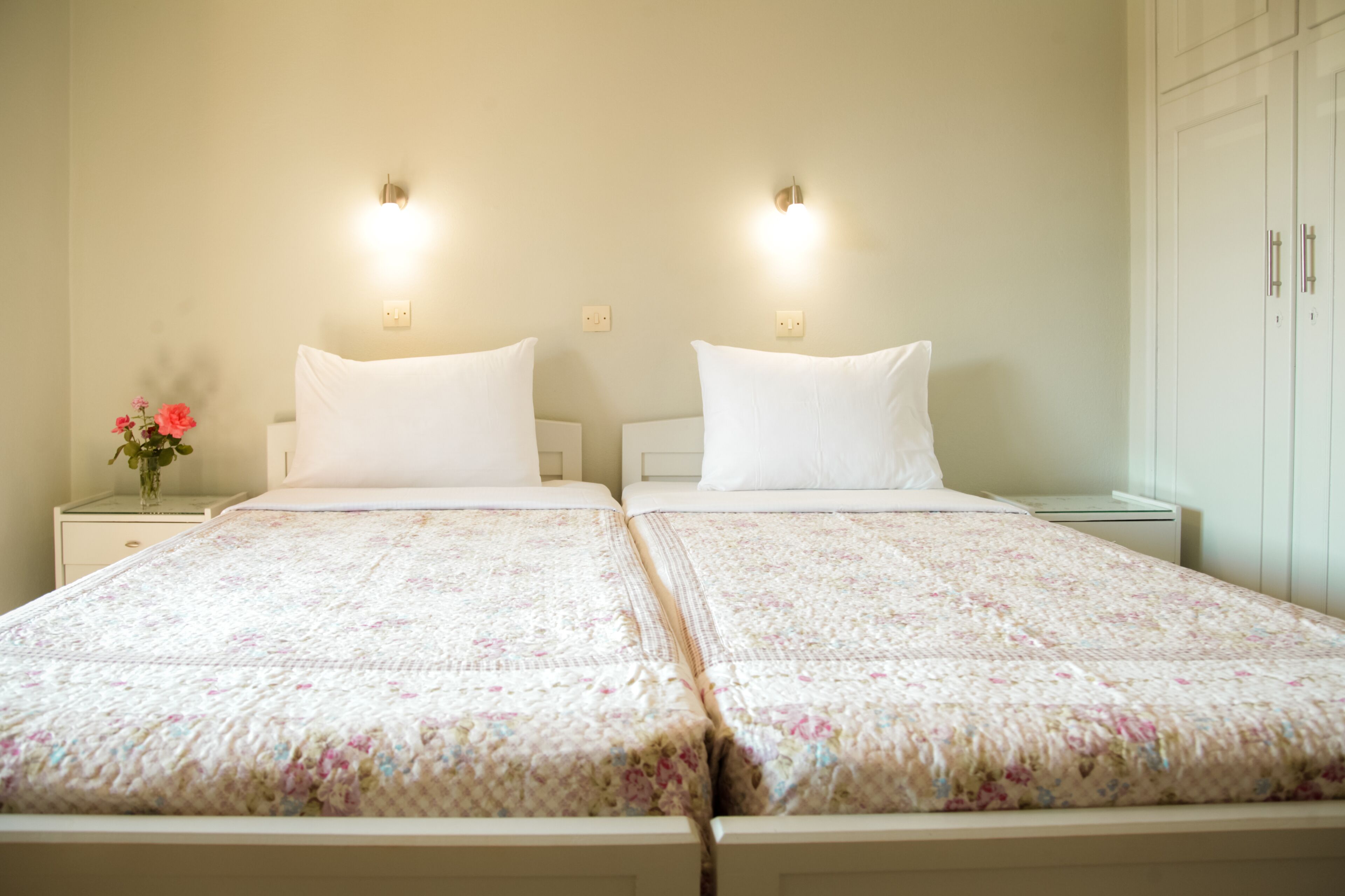 Una cama doble con colcha estampada, dos mesitas de noche, un florero con flores e iluminación en la pared.