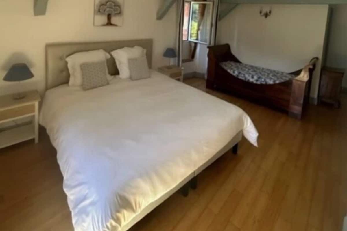 A bedroom with a large bed, a wooden floor, a nightstand, a bedside table, a window, and a wooden bench.