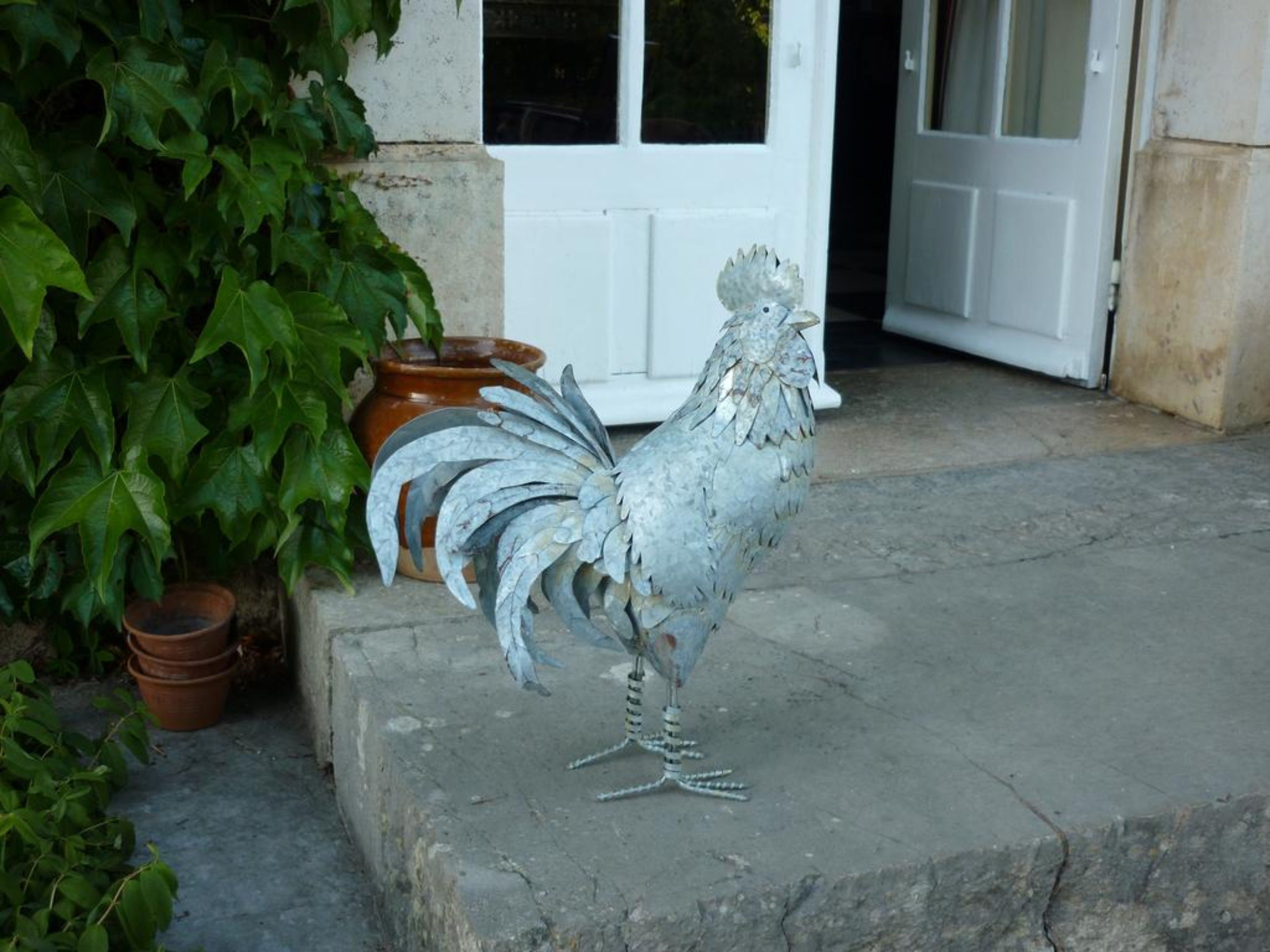 A metal rooster sculpture on a concrete step outside a building with a white door and potted plants nearby.