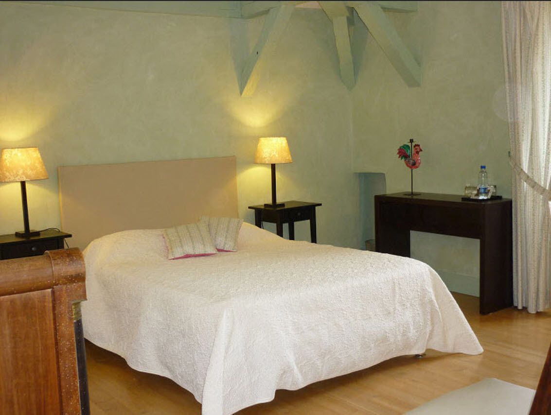 A neatly made bed with a white bedspread, two bedside lamps, a small table with a flower vase, and a wooden chair.