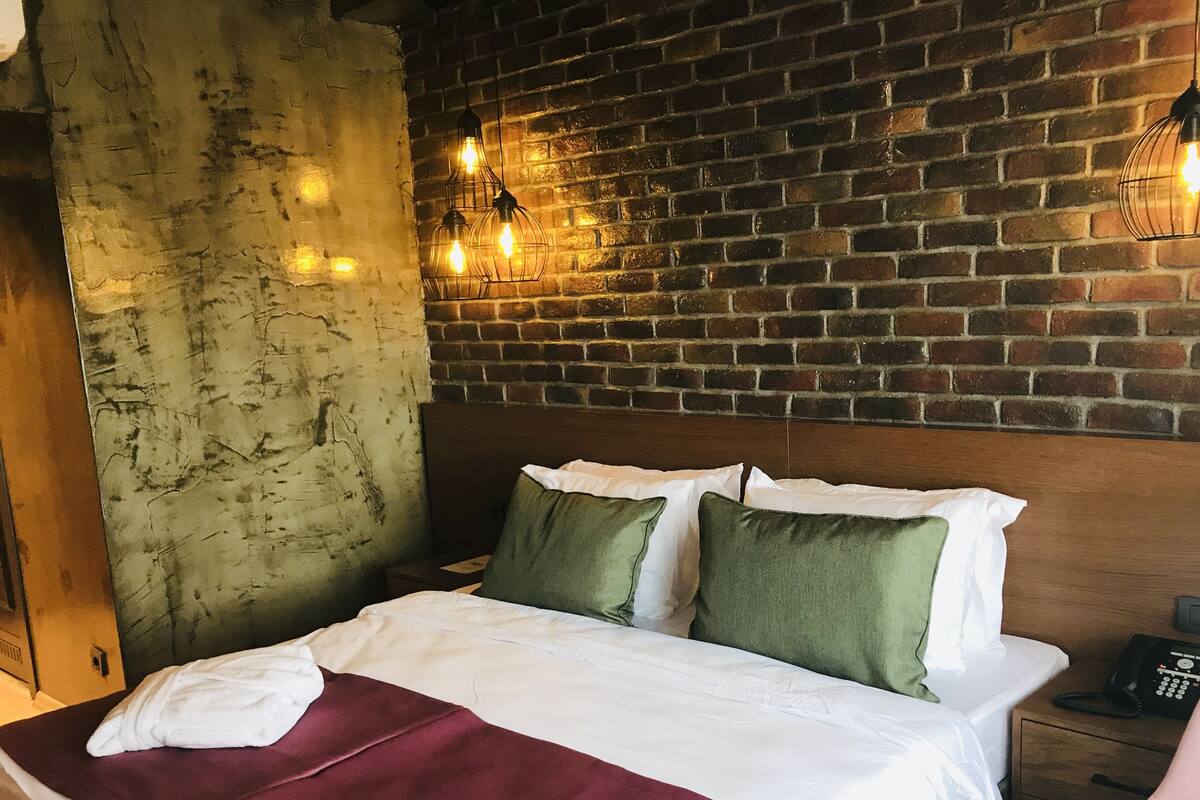 A neatly made bed with white and green pillows, a maroon blanket, and a wooden headboard against a brick wall.