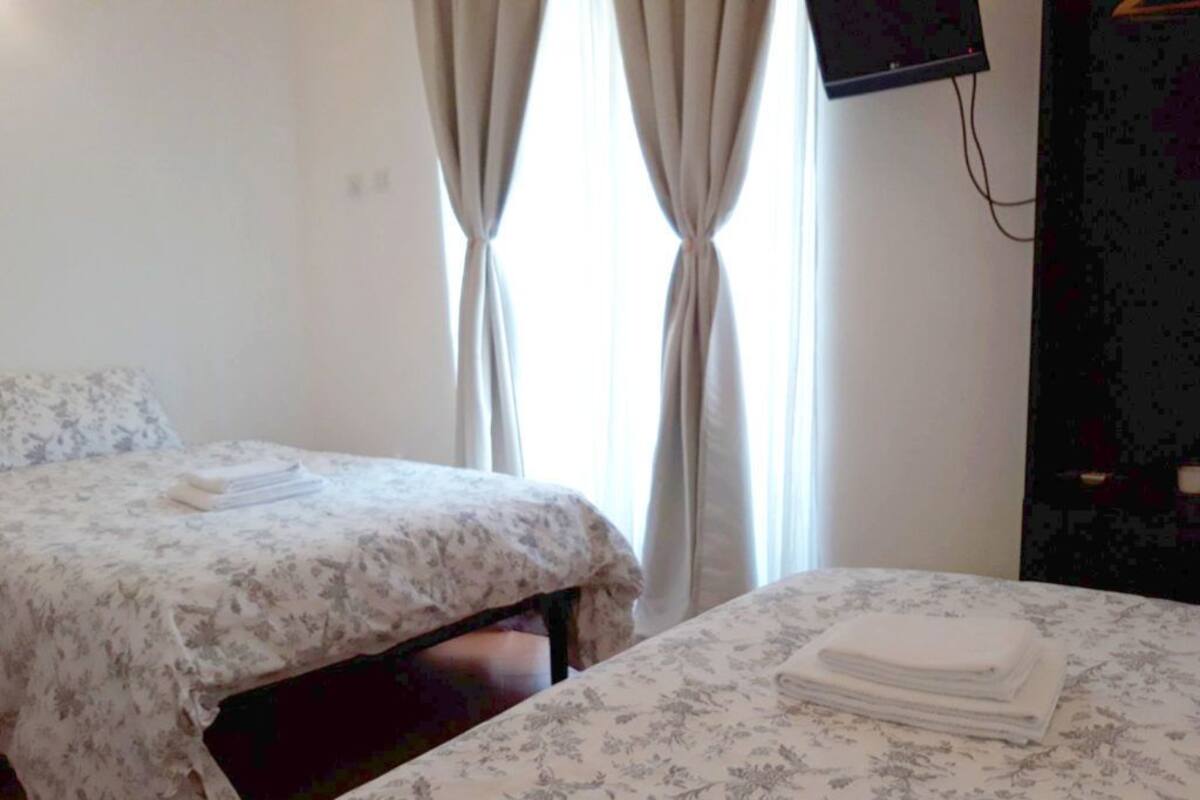 Habitación de hotel con dos camas, una con cubrecama floral y la otra con cubrecama lisa. Hay un televisor montado en la pared y una ventana con cortinas.