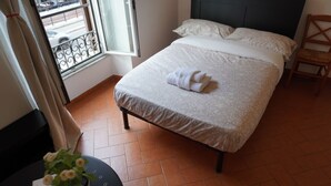 Een net opgemaakt bed met een gevouwen handdoek, een houten stoel, een klein tafeltje met een potplant en een raam met uitzicht op de straat.