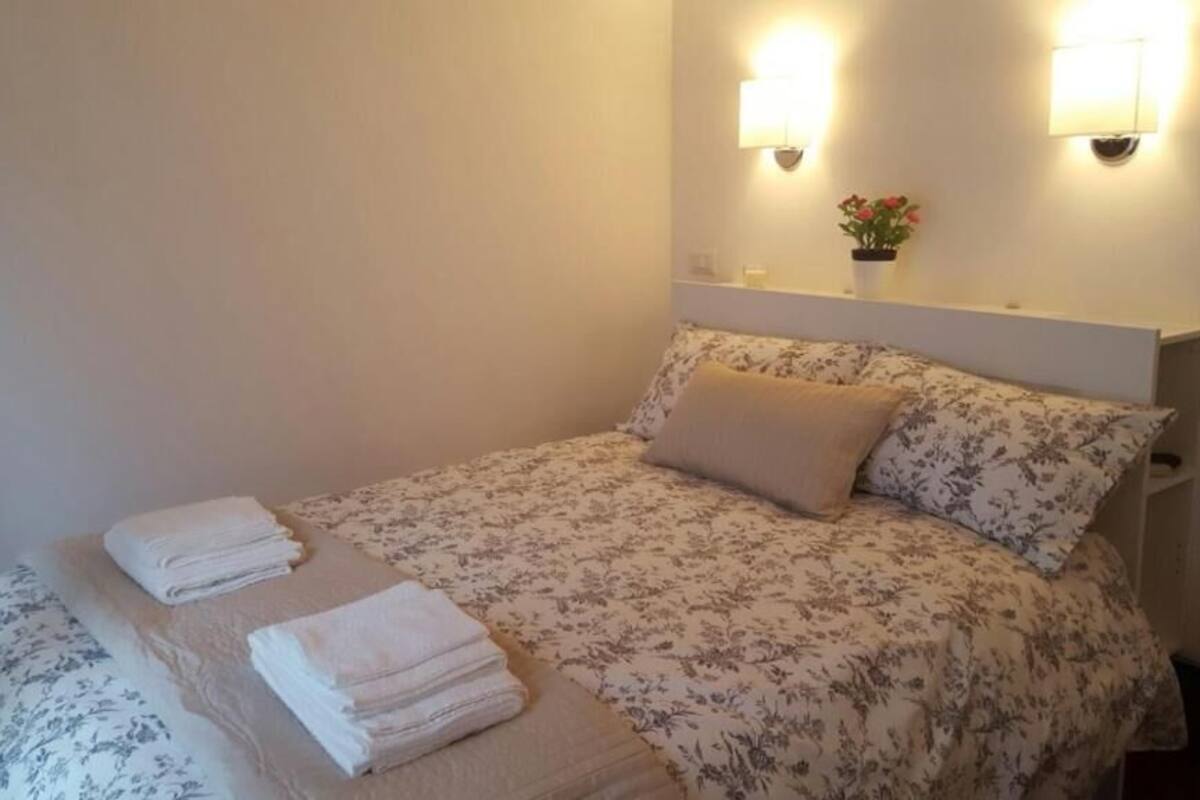 Una cama bien hecha con ropa de cama floral y dos almohadas, una mesita de noche con una lámpara y una pequeña planta en maceta.