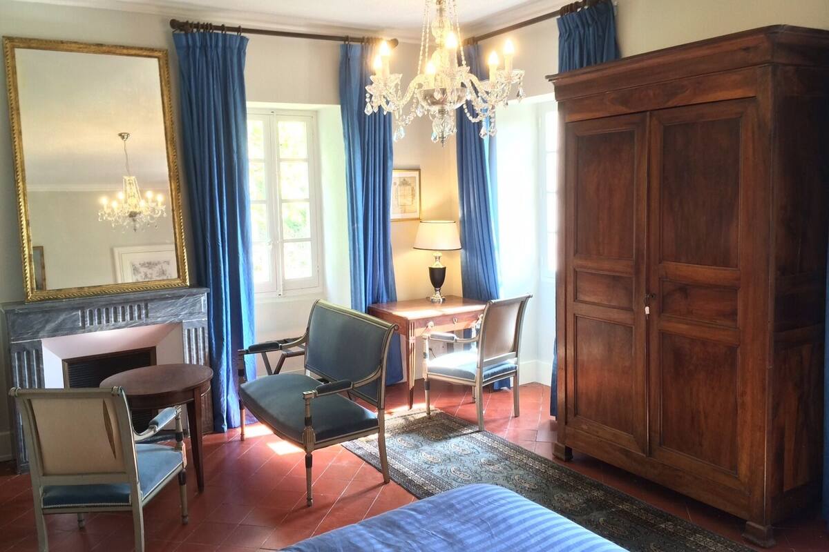 A room with a bed, two chairs, a wooden wardrobe, a desk with a lamp, a chandelier, and a large mirror.